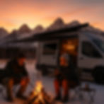 A couple in their 60s sits by a campfire in the snowy Colorado Rockies at sunset, enjoying coffee beside their custom Ford Transit camper van with the awning extended. The van glows warmly in the golden-hour light, surrounded by snow-covered trees and mountain peaks.